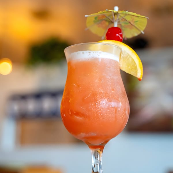 A vibrant orange cocktail in a tall glass, garnished with a lemon slice, cherry, and paper umbrella.