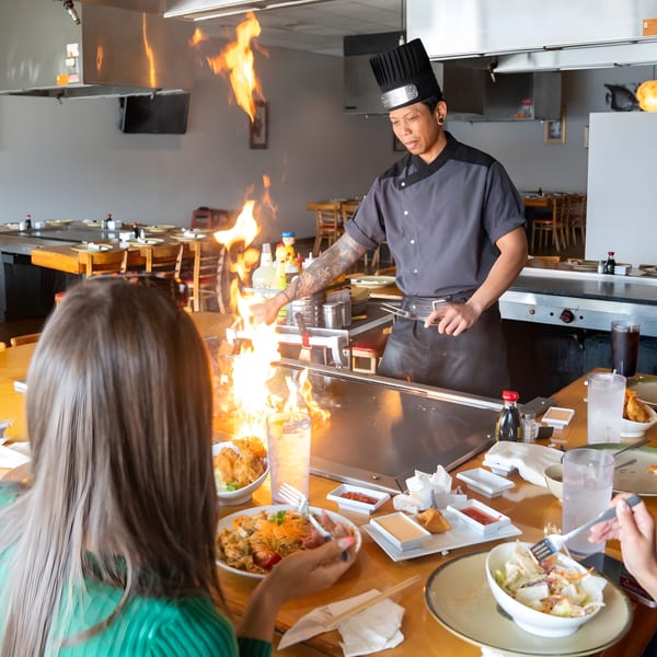 A hibachi chef cooks with large flames on a grill as customers watch and eat at Shiki Japanese Thai Sushi Bar.