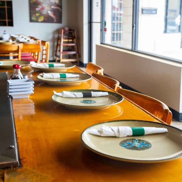 A long wooden hibachi table with place settings, soy sauce, and chairs in a Japanese restaurant.