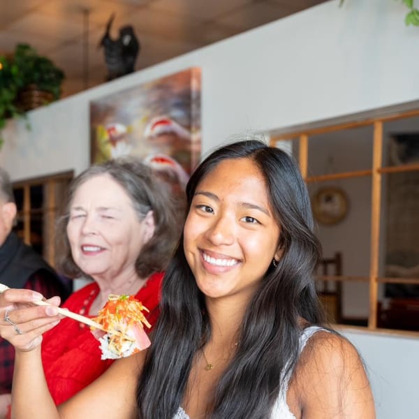 A smiling young woman holds a piece of sushi with chopsticks at Shiki Japanese Thai Sushi Bar.