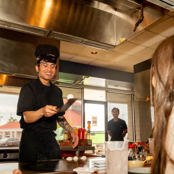 A smiling Shiki Japanese Thai Sushi Bar chef flips an egg on a hibachi grill for customers.